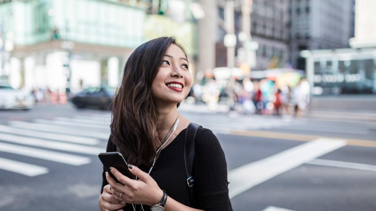 一个微笑的女人在大城市过马路，手里拿着智能手机，戴着耳塞.